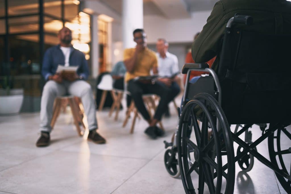 Au premier plan on voit un salarié en fauteuil roulant et en fond, de manière floutée, d'autres salariés assis sur une chaise