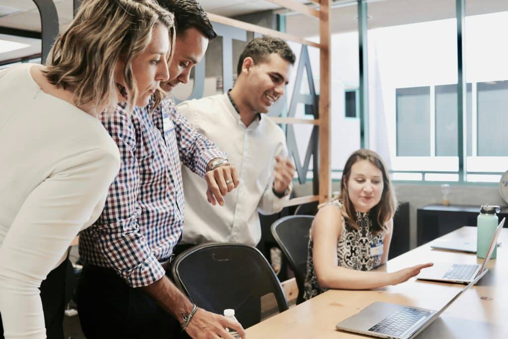 Quatre collègues échangent autour de plusieurs ordinateurs portables dans un espace de travail partagé dont qui est en train de regarder sa montre