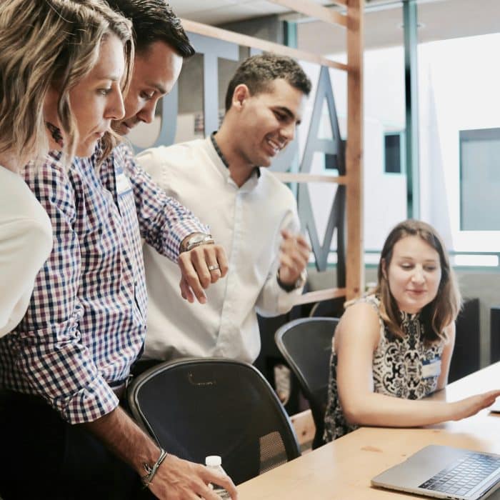 Quatre collègues échangent autour de plusieurs ordinateurs portables dans un espace de travail partagé dont qui est en train de regarder sa montre